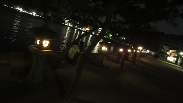 a line of lit stone lanterns along the shoreline with city lights in the far distance